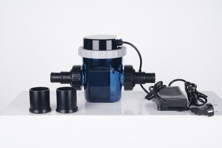 Gerador de Cloro NT Pro da Netuno posicionado sobre uma superfície branca, com corpo transparente em tom azul, conexões laterais para encanamento, tampa superior preta e branca, dois adaptadores ao lado e fonte de alimentação com cabo elétrico dispostos à direita. Fundo neutro e iluminação clara destacando os detalhes do equipamento.